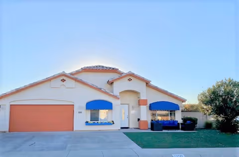 Front exterior view of a single-story residential-style building with a two-car garage, two windows with blue awnings, a white front door, and a small lawn area with outdoor seating on the right side under a clear blue sky.