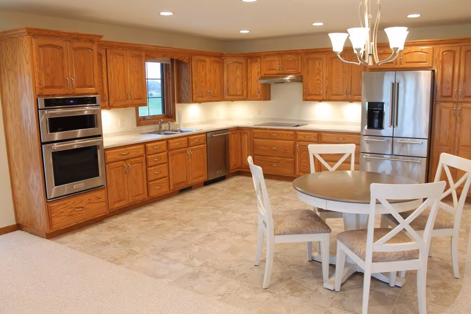A spacious kitchen with wooden cabinets, stainless steel double oven, dishwasher, and refrigerator. There is a round dining table with four white chairs in the foreground. The floor is tiled and there is a window above the sink.