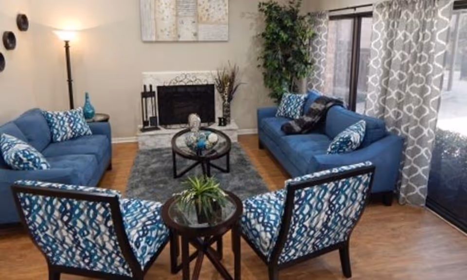 Bright living room with blue sofas and patterned chairs arranged around a round coffee table in front of a fireplace and sliding glass doors.