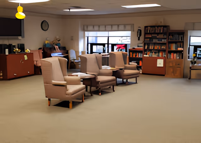 A spacious room with beige carpet and several beige armchairs arranged in a row facing the camera. In the background, there are bookshelves filled with books, a piano, a clock on the wall, and large windows letting in natural light. The room has a calm and cozy atmosphere, likely a common area in a senior living facility.