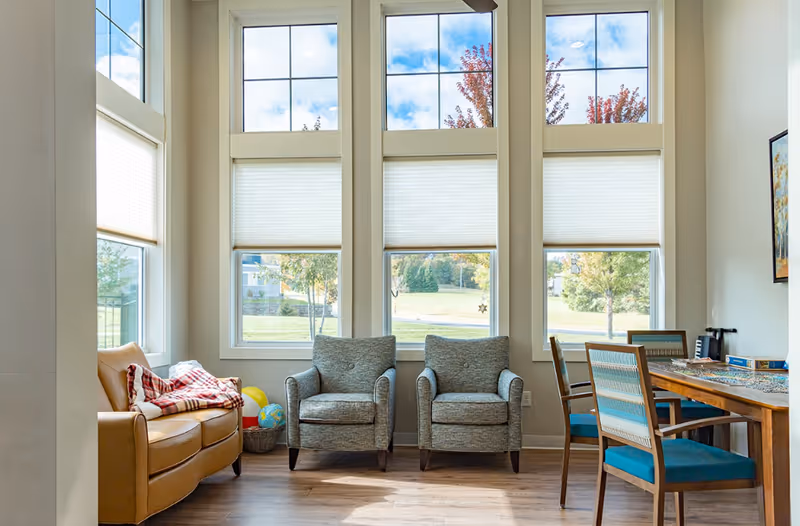 Sunlit communal living area with tall windows, a leather sofa, two upholstered armchairs, and a wooden dining table with chairs.