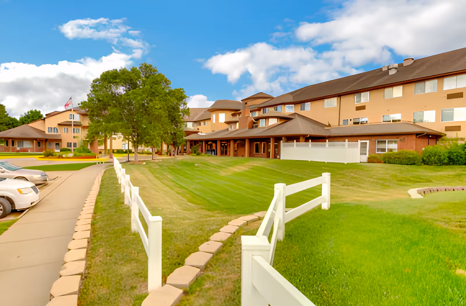 Exterior view of a large senior living facility building with beige walls and brown roof, surrounded by green grass, trees, and a white wooden fence. Several parked cars are visible on the left side along a paved driveway under a partly cloudy blue sky.