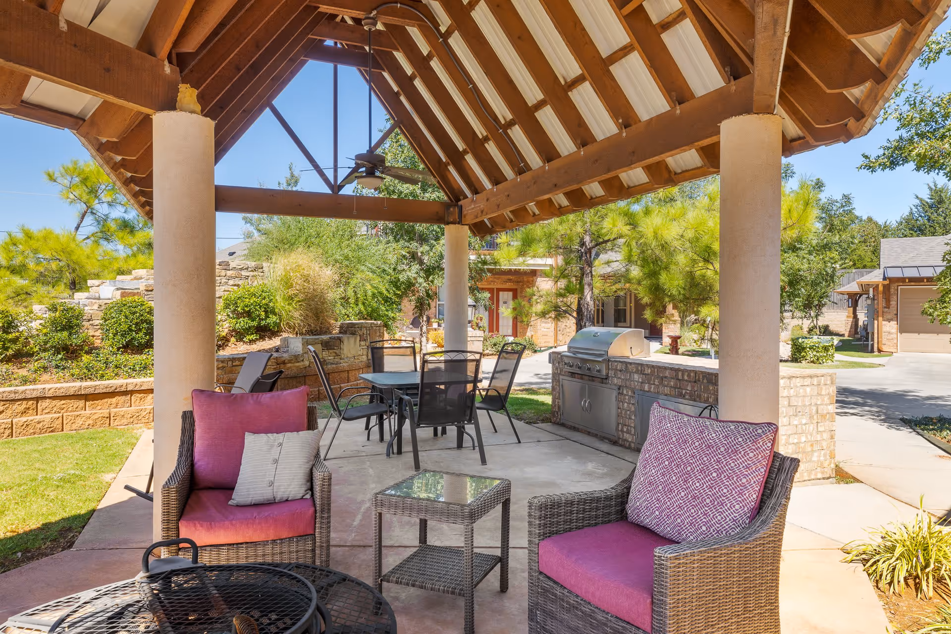 Outdoor covered patio area with a wooden roof supported by columns, featuring wicker chairs with purple cushions, a small glass-top table, a dining table with black chairs, and a built-in brick grill. Surrounding the patio are green trees, shrubs, and a clear blue sky.