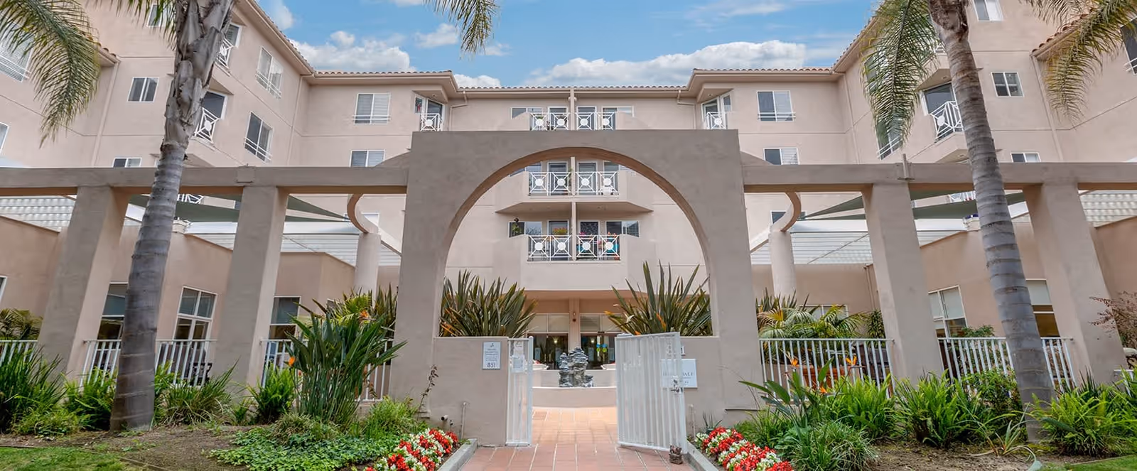 Exterior view of Brookdale Santa Monica Gardens, showing a multi-story beige building with balconies, palm trees, and a gated entrance with an archway surrounded by landscaped greenery and flowers under a partly cloudy sky.