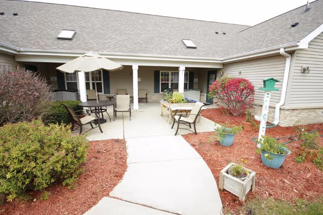 Outdoor patio area at Brookdale Delta with a concrete walkway leading to a covered seating area. The patio features tables with umbrellas, chairs, raised garden beds, and various potted plants surrounded by red mulch. The building exterior is beige with a sloped roof and skylights.