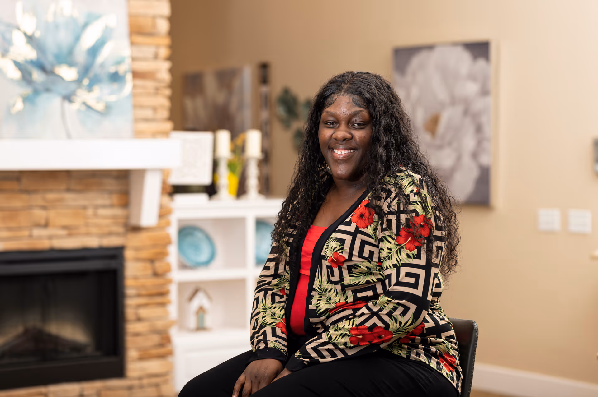 A smiling woman with long curly hair wearing a patterned jacket with red flowers sits on a chair in a cozy living room. Behind her is a stone fireplace, white shelving with decorative items, and wall art featuring floral designs.