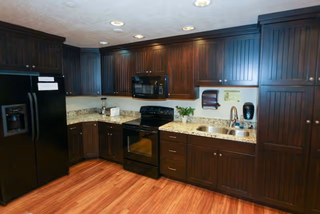 A kitchen area with dark wooden cabinets, a black refrigerator, black stove with oven, black microwave, granite countertops, a stainless steel sink, and wooden flooring. There is a paper towel dispenser and a soap dispenser mounted on the wall above the sink, along with a small plant on the counter.