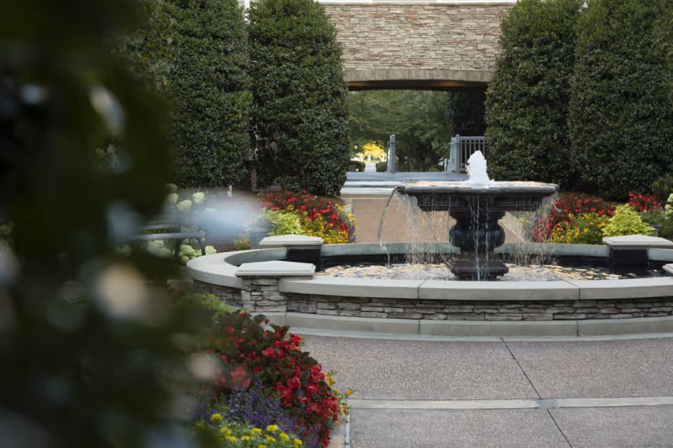 A stone water fountain surrounded by colorful flower beds and neatly trimmed bushes in an outdoor garden area with a stone archway in the background.