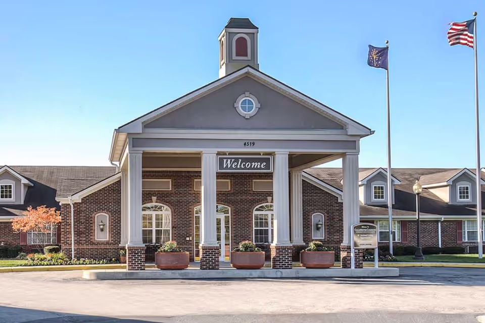 Front entrance of a brick senior living building with a columned portico, a 'Welcome' sign and flagpoles.