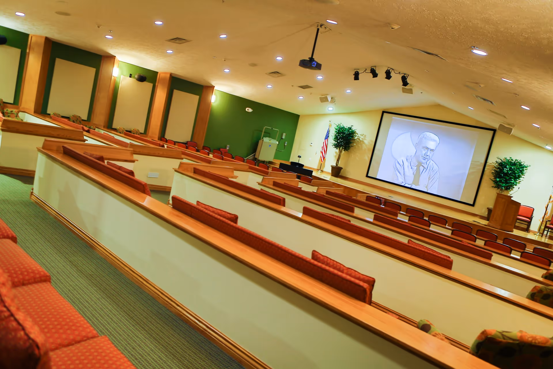 Interior view of a lecture or presentation room with tiered seating featuring red cushioned benches. A large screen at the front displays a black and white image of a man. The room has green and beige walls, ceiling lights, and an American flag near the screen. There are also two potted plants flanking the screen.