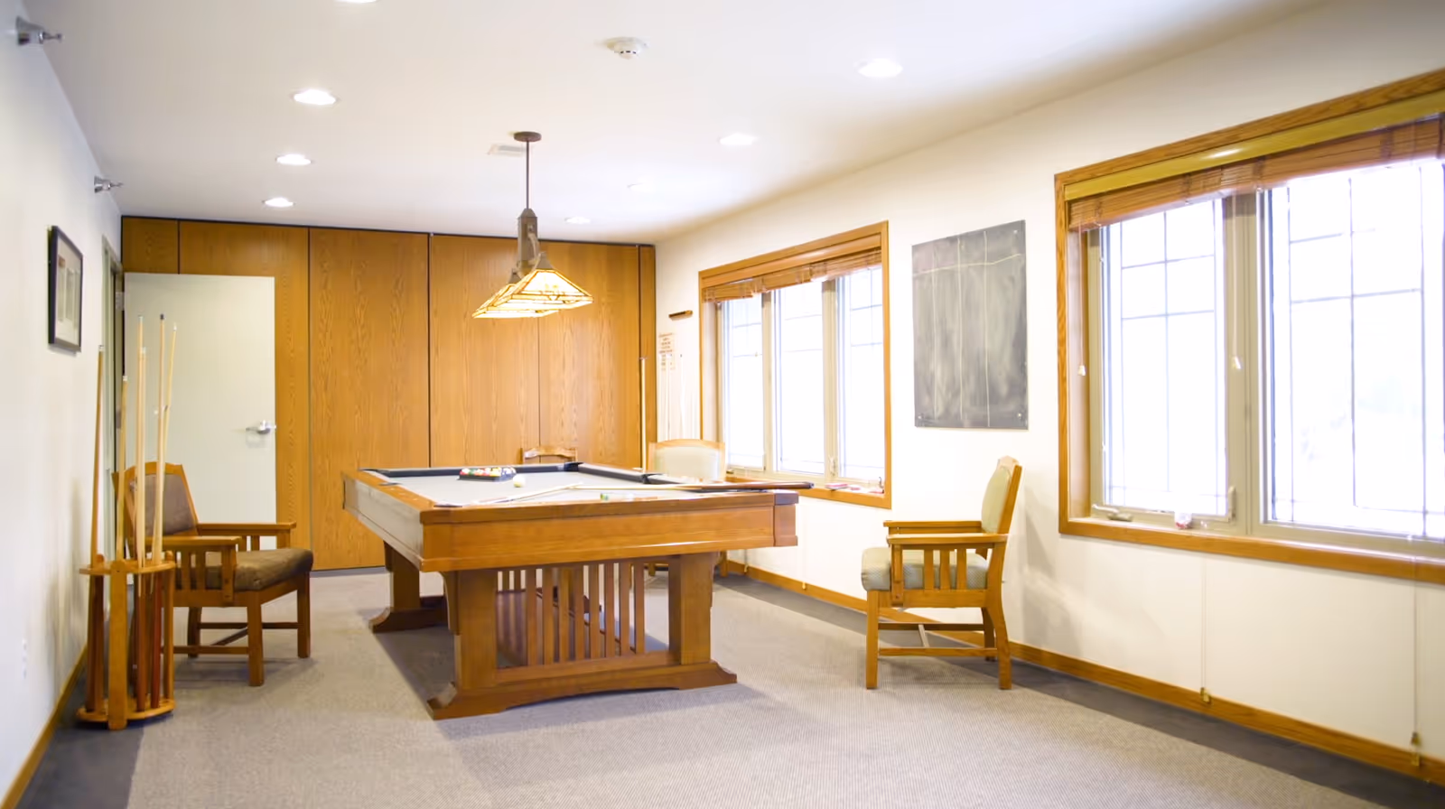A bright room with a pool table in the center, surrounded by wooden chairs with cushions. The room has large windows with wooden blinds, a hanging light fixture above the pool table, and a rack holding pool cues. The walls are light-colored with wooden trim and a chalkboard on one wall.