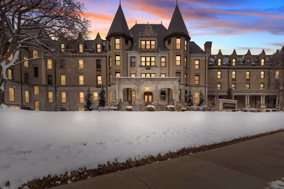 Snow-covered lawn in front of a large historic multi-story brick building with turrets and lit windows at dusk.