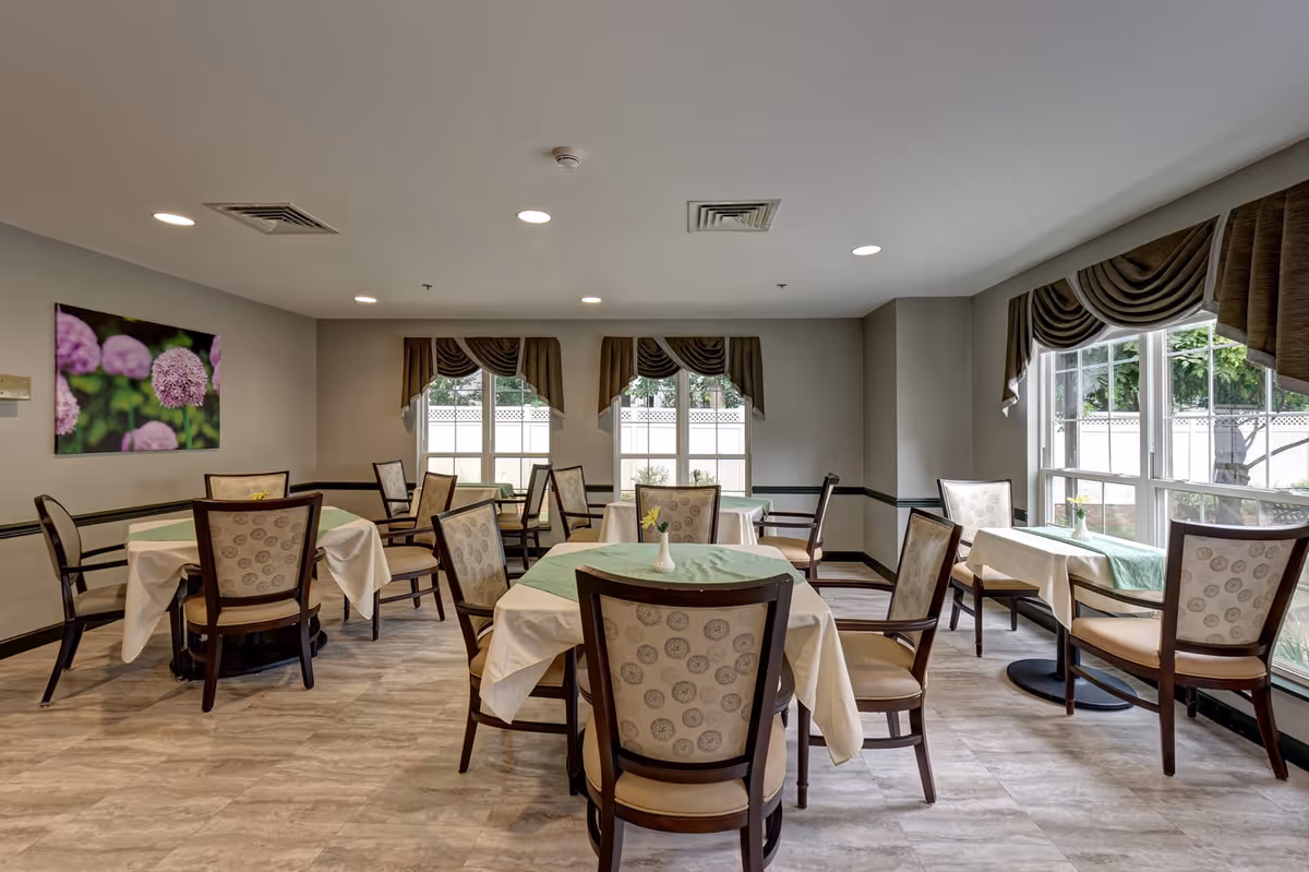 A dining room with several tables covered with white tablecloths and green runners. Each table is surrounded by wooden chairs with patterned upholstery. Large windows with brown valances let in natural light, and a floral painting hangs on the wall. The floor has a light wood-like finish.