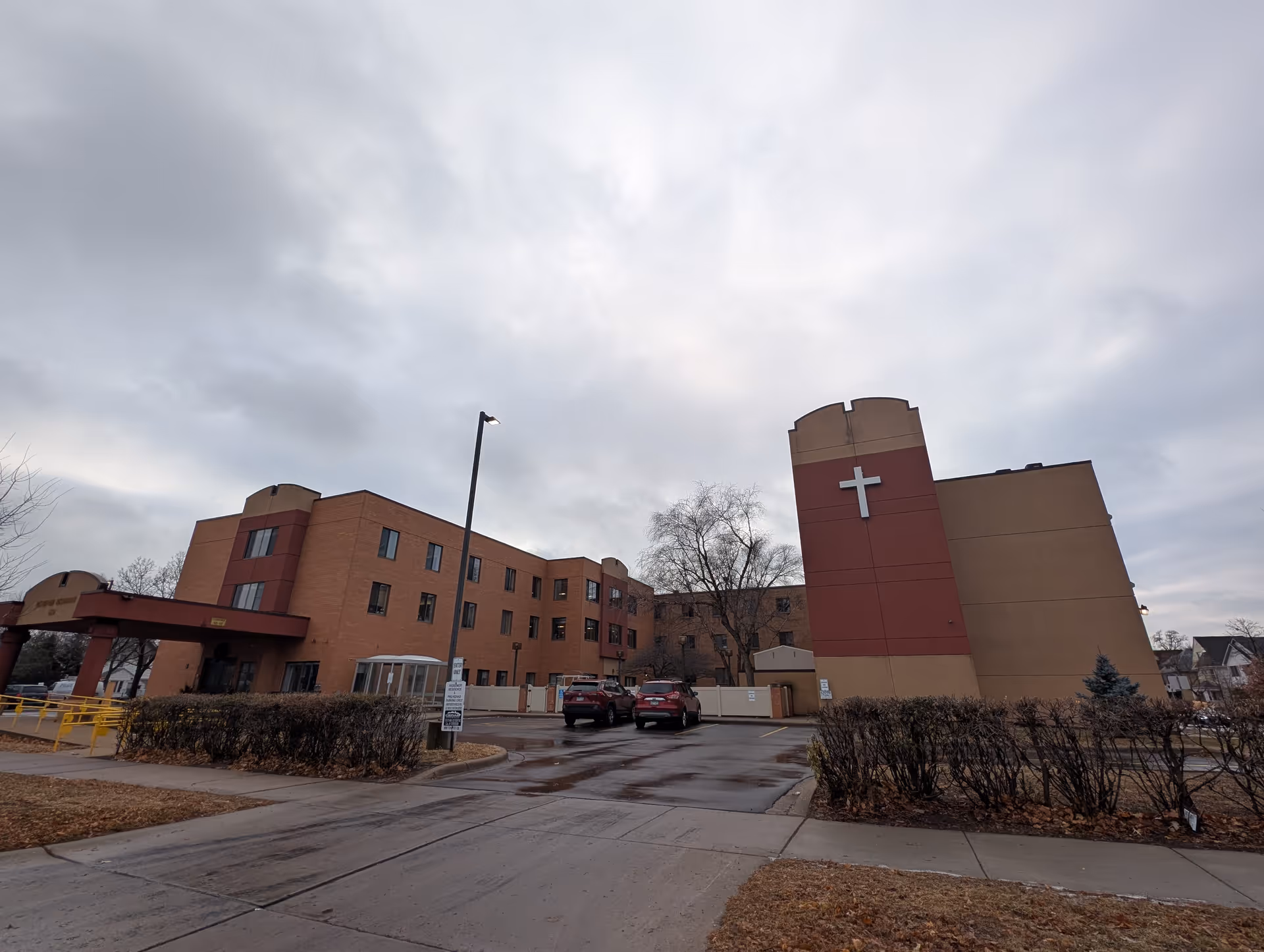 Exterior view of a three-story senior living facility building with a large cross mounted on a red and beige section of the building. There are two cars parked in front, leafless bushes, and a cloudy sky overhead.