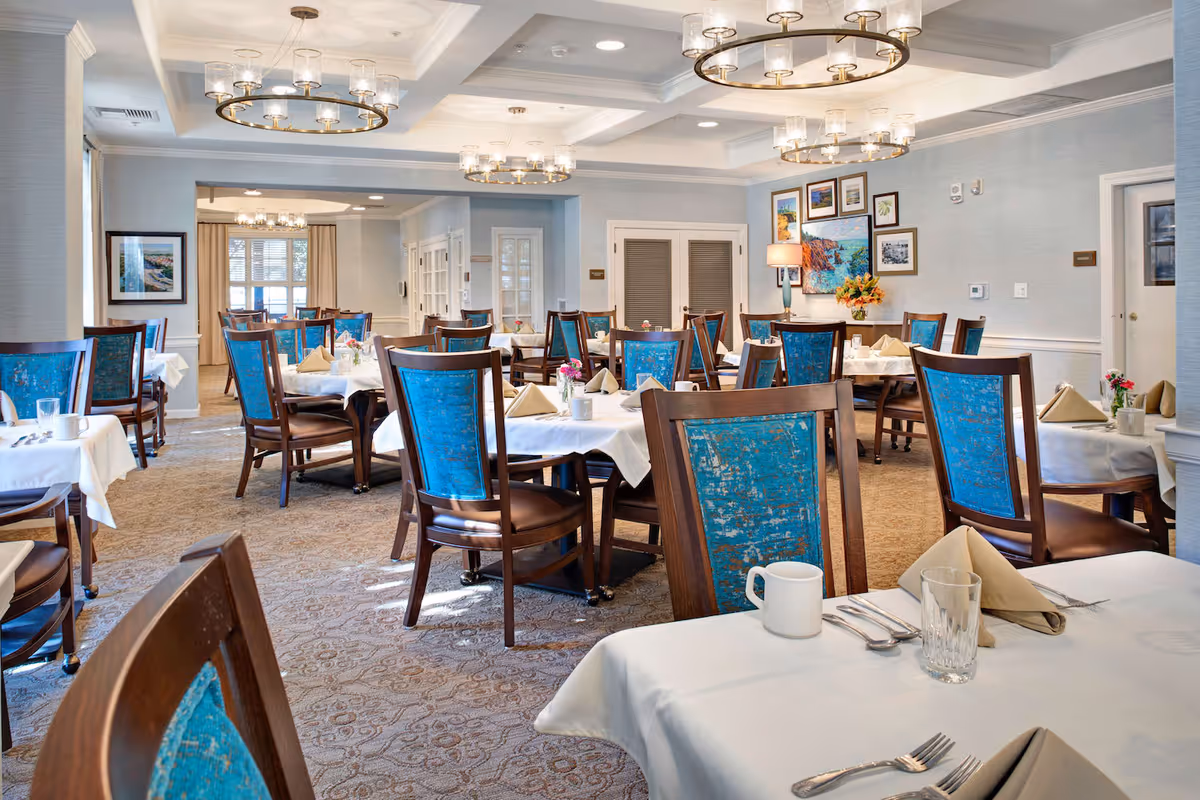 A bright and elegant dining room with multiple tables covered in white tablecloths, each set with beige napkins, glassware, and silverware. The chairs have wooden frames with blue upholstered backs. The room features patterned carpet, soft blue walls, framed artwork, and modern circular chandeliers hanging from the ceiling.