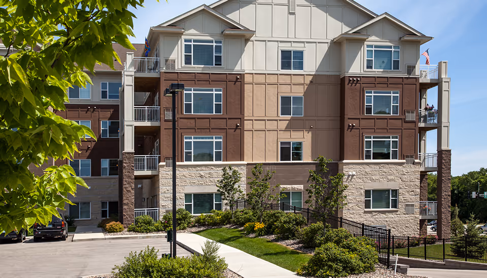 Exterior view of a multi-story senior living facility building with beige, brown, and stone facade. There are several windows, balconies, a parking area with cars, and landscaped greenery including bushes and small trees along a sidewalk.