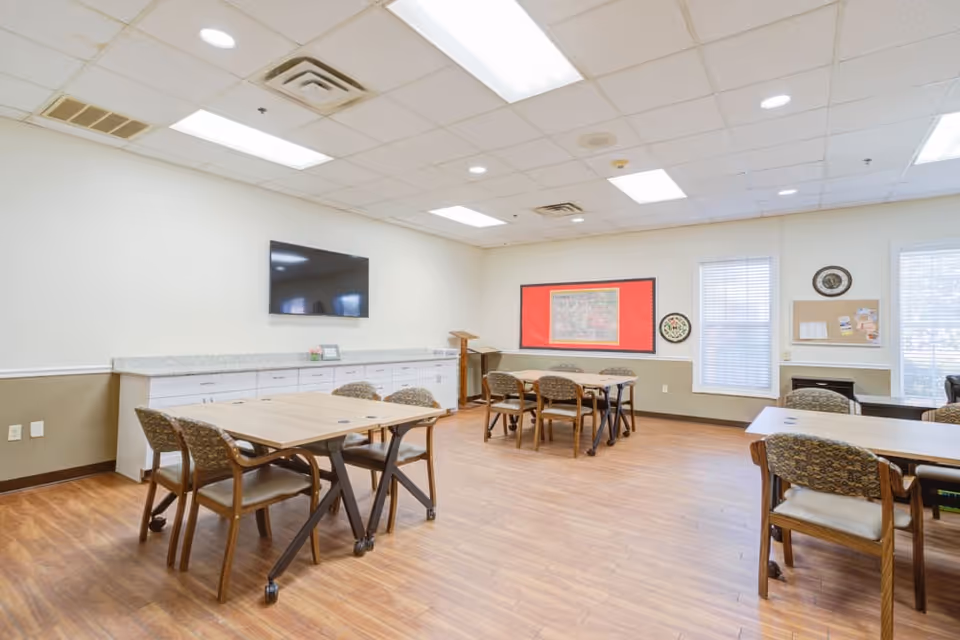 A bright, spacious room with wooden floors and several tables with chairs arranged around them. The walls are light-colored with a darker wainscot. A flat-screen TV is mounted on one wall above a long counter with cabinets. There are windows with blinds letting in natural light, a bulletin board with a red border, and some wall decorations.