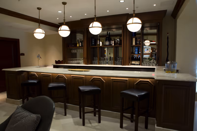 A well-lit bar area with a marble countertop and four black cushioned bar stools. Behind the bar is a wooden cabinet with glass shelves holding bottles and glasses. Four round pendant lights hang from the ceiling above the bar. A chair with a cushion is partially visible in the foreground.