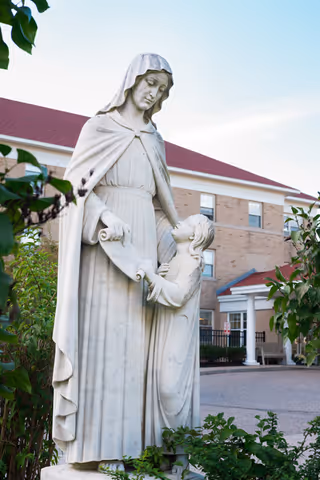 White stone statue of a robed woman holding a scroll and a child, set in front of a brick nursing center building.