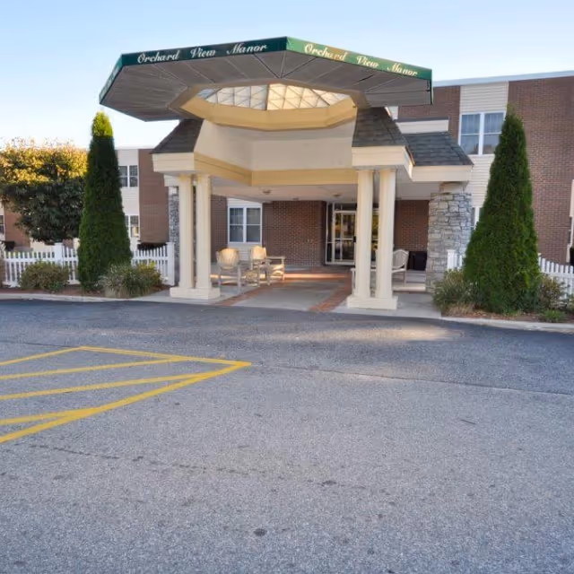 Entrance of Orchard View Manor featuring a covered drop-off area with white columns, two chairs, and a brick building facade with windows. There are two tall evergreen shrubs on either side and a white picket fence in the background.