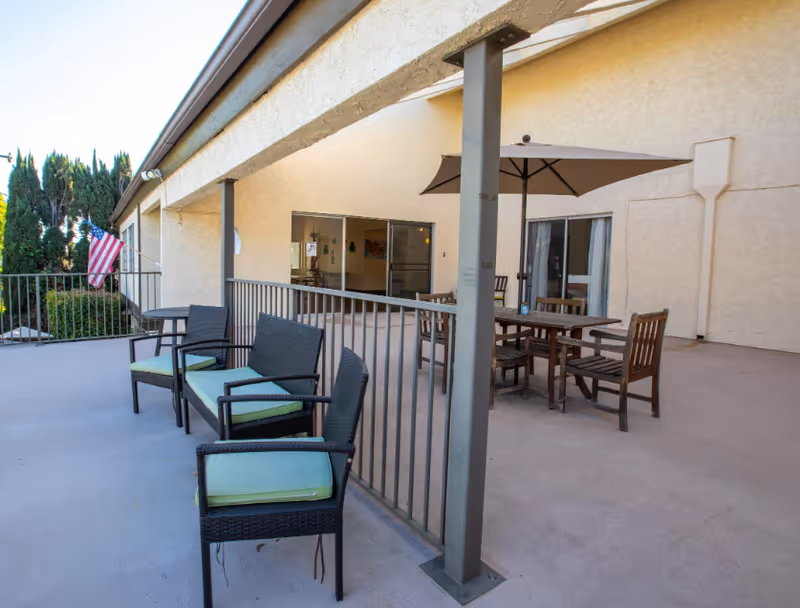 Outdoor patio area at Whittier Glen Assisted Living with several chairs and tables, including a table with an umbrella. The patio is adjacent to the building with sliding glass doors and an American flag is visible in the background.