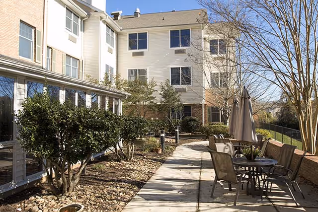 Outdoor patio area at The Parker with a paved walkway, outdoor table and chairs with an umbrella, surrounded by bushes and trees, adjacent to a multi-story building with large windows.