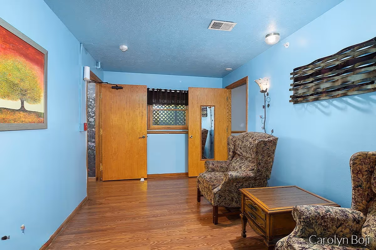 A small sitting area with two patterned armchairs and a wooden side table between them. The walls are painted light blue and decorated with a metal wall art piece and a painting of a tree. There are two wooden doors, one with a window, and a window with a dark valance in the background. The floor is wood laminate.