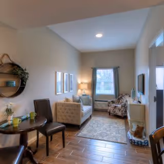 A cozy living room area with a small round dining table and two chairs on the left, a beige tufted sofa, a patterned armchair near a window with curtains, a floor lamp, a rug on wooden flooring, and decorative wall art and plants.