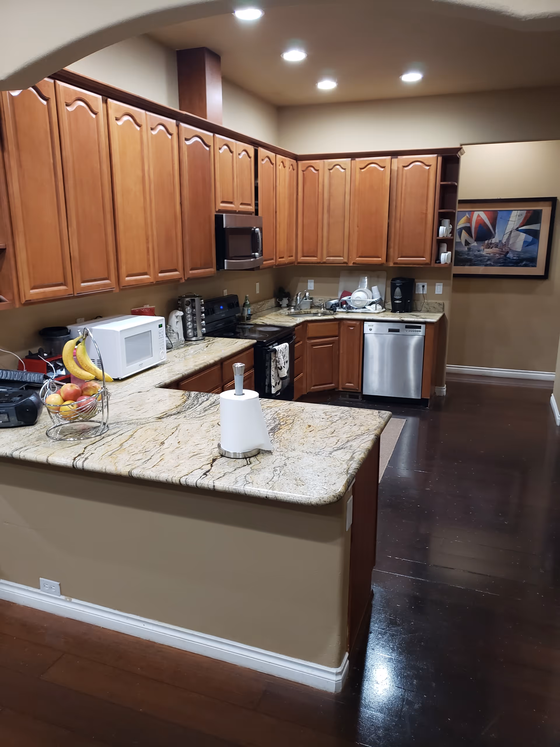 A kitchen with wooden cabinets, granite countertops, and dark wooden flooring. The kitchen includes a microwave, stove, dishwasher, coffee maker, and a fruit basket with bananas and apples on the counter. There is a paper towel holder on the countertop and a framed picture of sailboats on the wall.