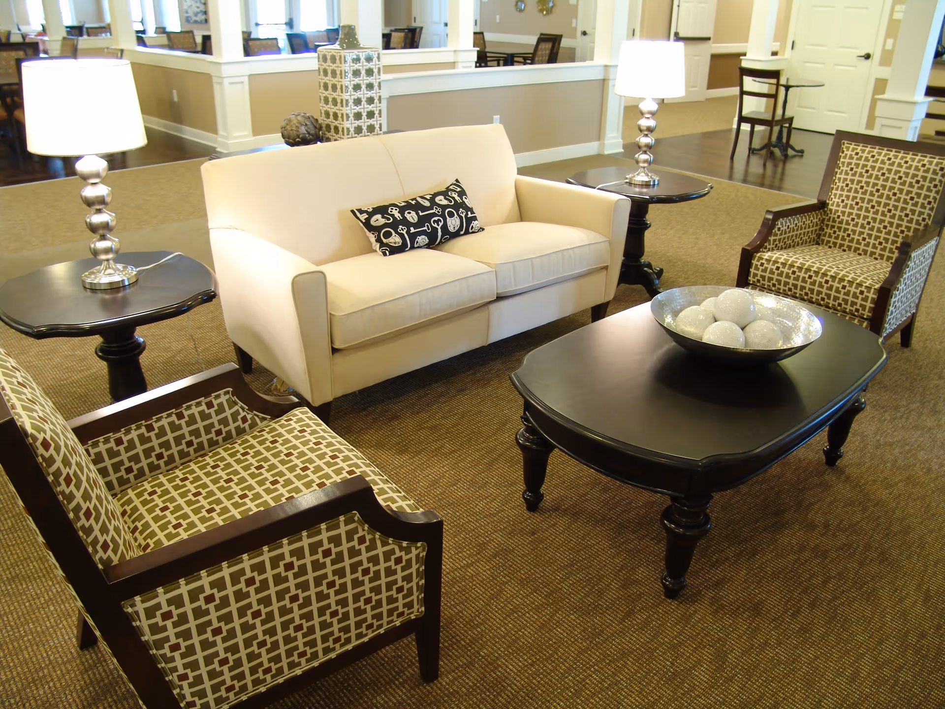 A cozy living room area with a beige loveseat, two patterned armchairs, a dark wooden coffee table with a decorative bowl of white spheres, and two side tables each with a silver lamp. The room has carpeted flooring and light-colored walls with a large mirror and windows in the background.
