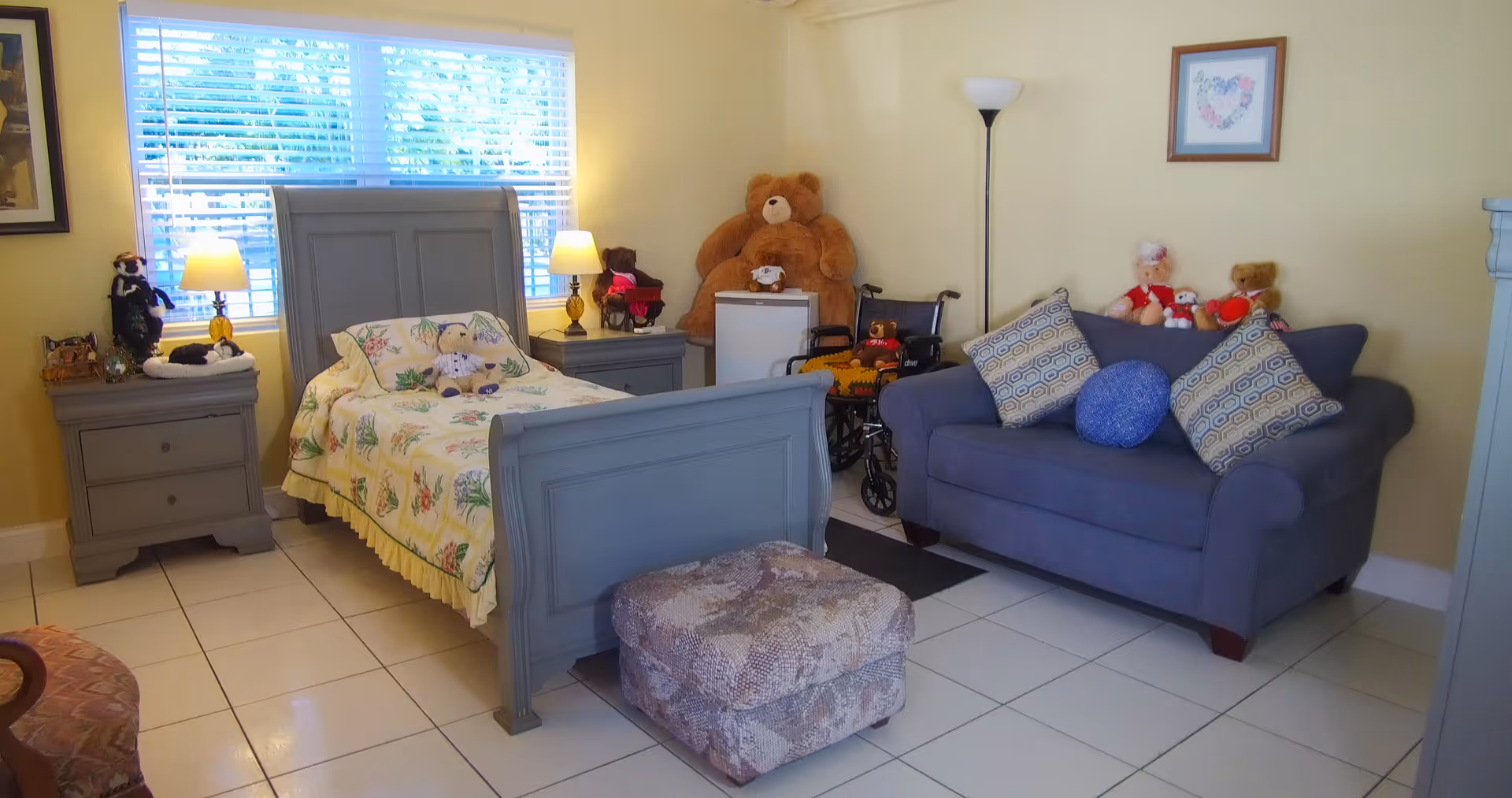 A cozy bedroom with a gray wooden bed featuring floral bedding and stuffed animals on it. There are two matching gray nightstands with lamps on either side of the bed. A large teddy bear and a wheelchair are positioned near a small white refrigerator. A blue loveseat with patterned pillows and more stuffed animals is against the wall. The room has tiled floors, a window with blinds, and soft yellow walls.