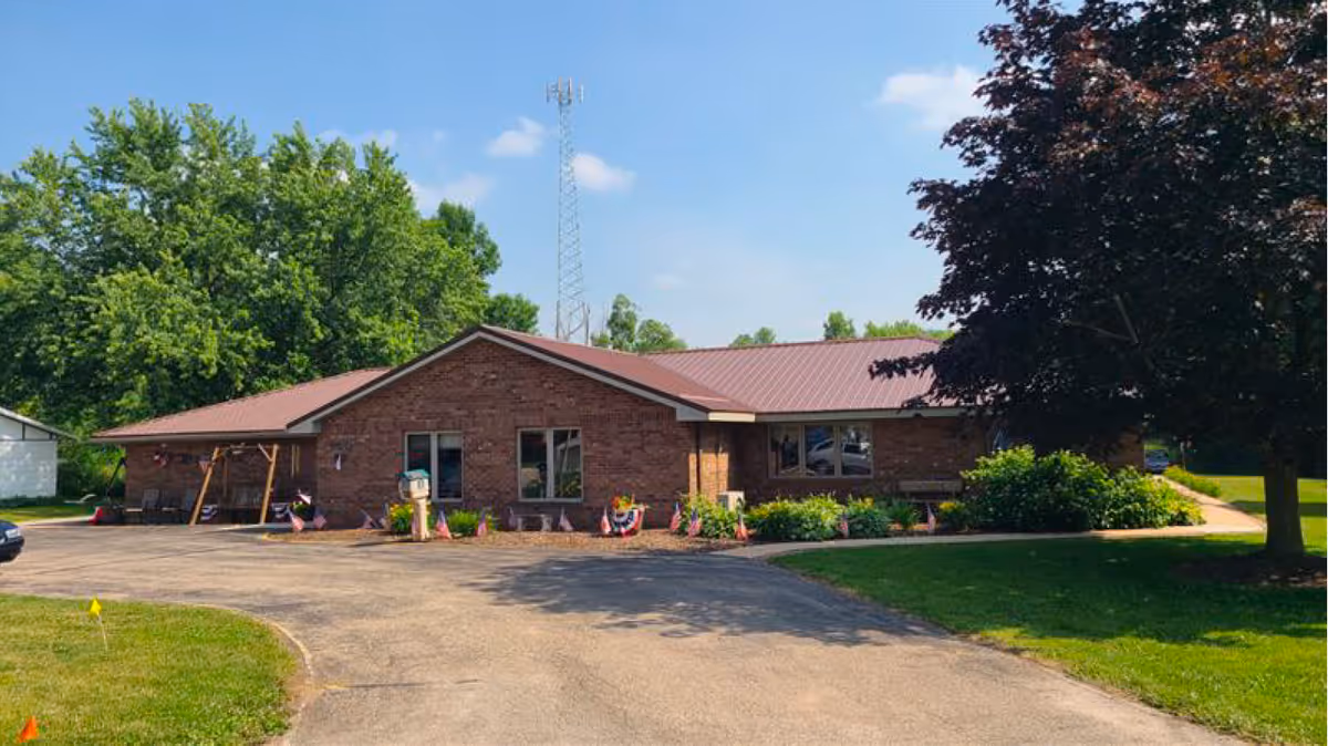 Single-story brick building with a red metal roof surrounded by greenery and trees, with a paved driveway and small garden beds in front, under a clear blue sky.