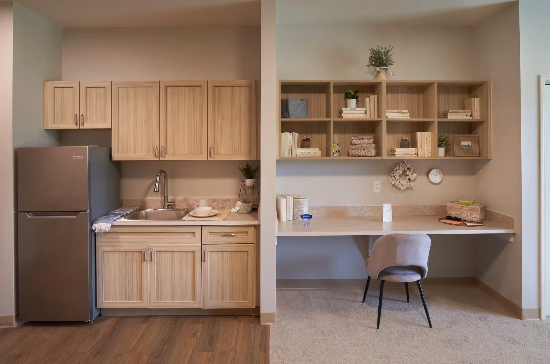 A small kitchenette area with light wood cabinets, a stainless steel refrigerator, and a sink with a faucet. Adjacent to the kitchenette is a built-in desk with open shelving above, decorated with books, plants, and small decorative items. A gray upholstered chair is placed at the desk. The floor transitions from wood in the kitchenette area to carpet in the desk area.