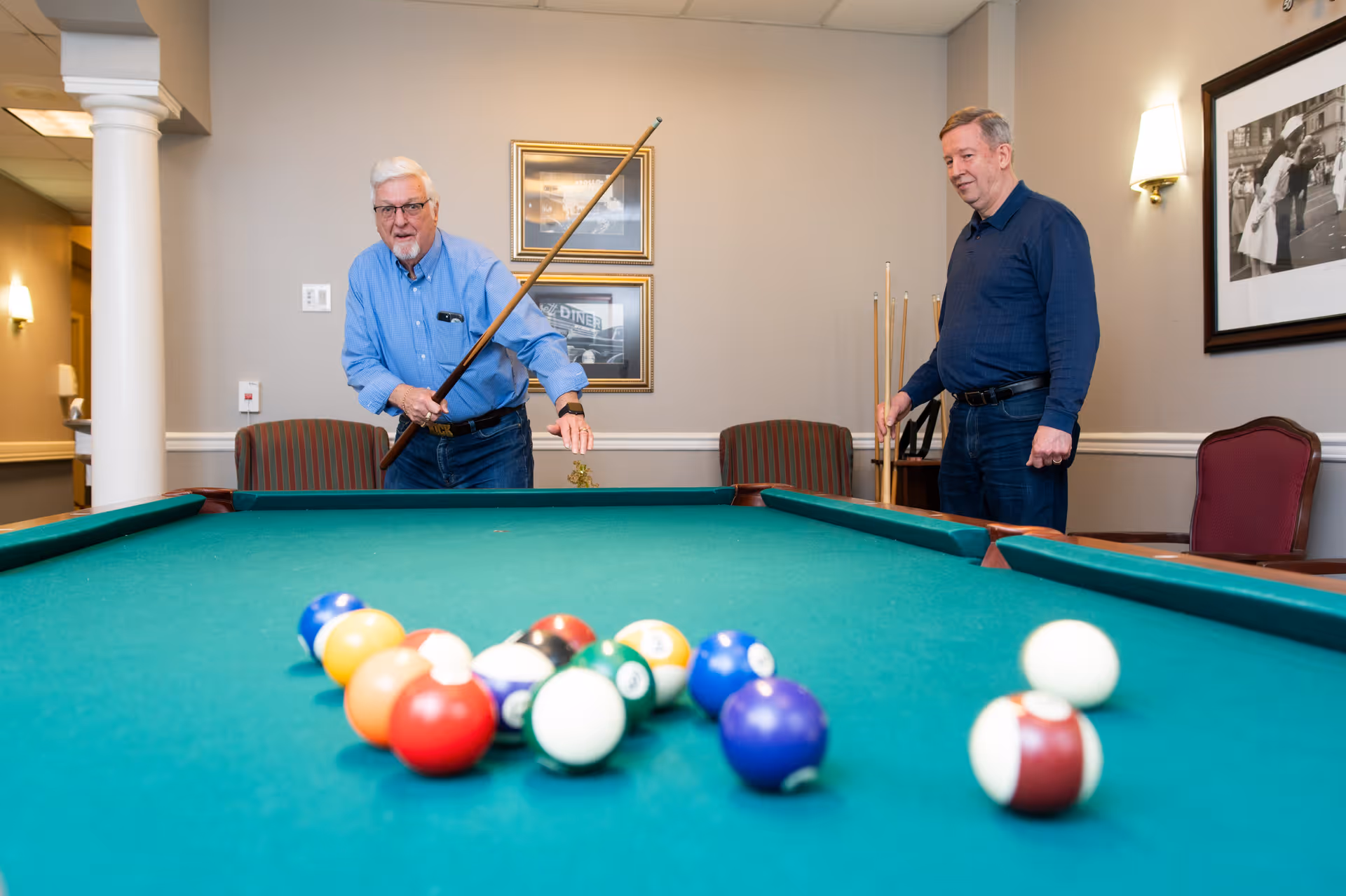 Two men stand by a pool table in a community recreation room with billiard balls racked in the foreground.