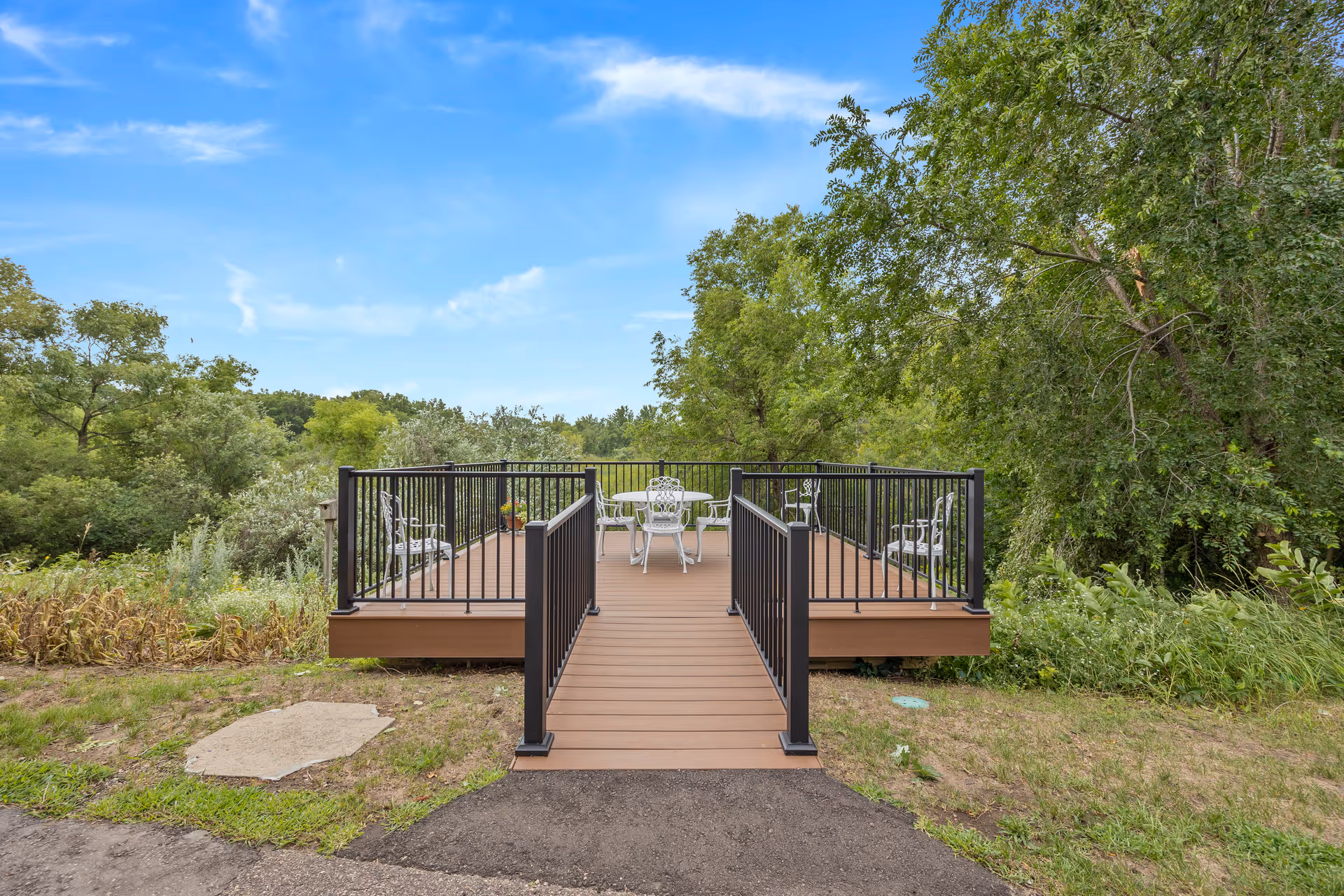 A wooden outdoor deck with black metal railings extending over a grassy area, surrounded by green trees and shrubs under a blue sky with some clouds. The deck has white metal chairs and a table arranged for seating.