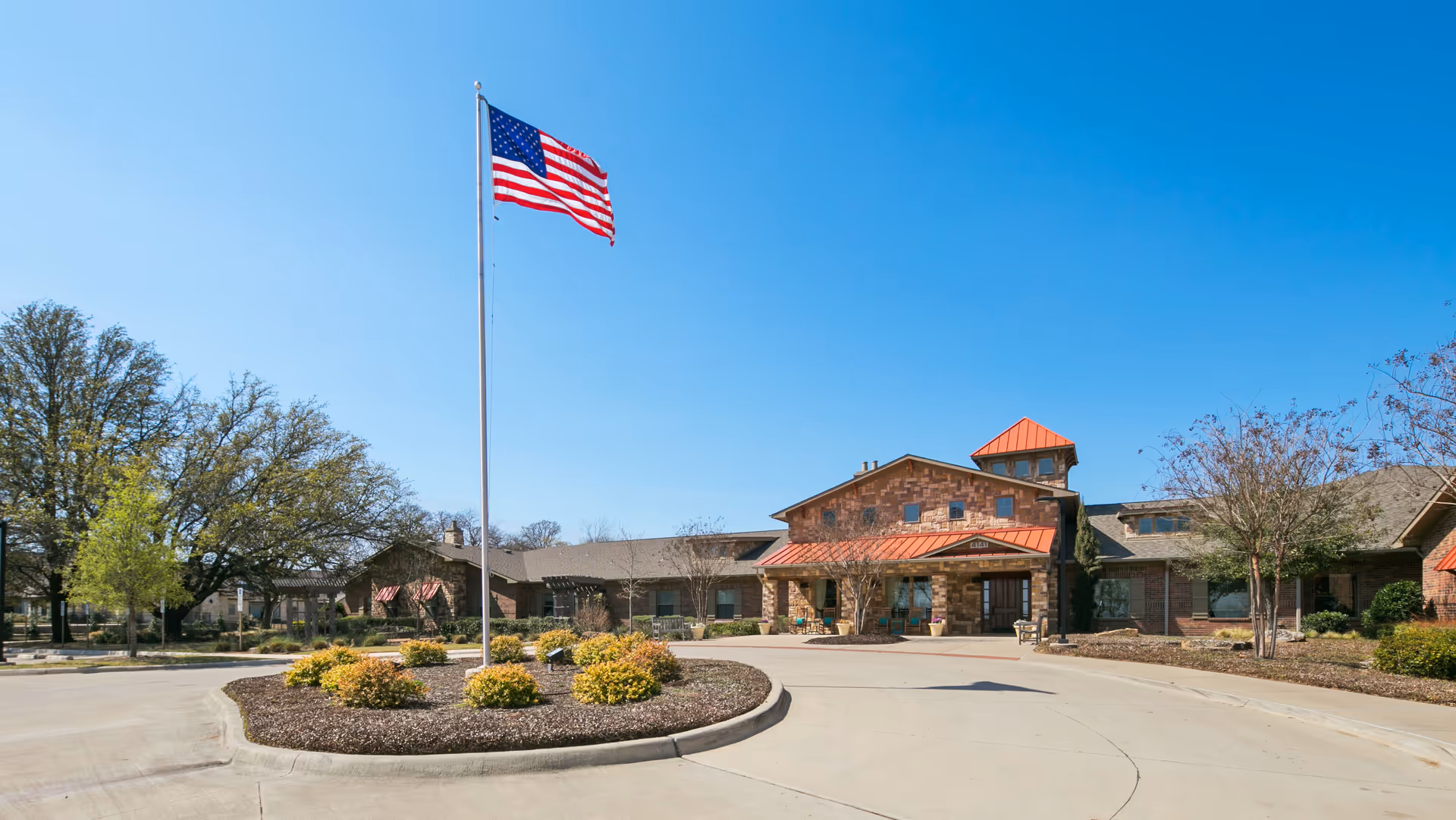 Exterior view of a single-story assisted living facility with a stone facade and red metal roof accents. A tall flagpole with an American flag is prominently displayed in front of the building, surrounded by landscaped bushes and trees under a clear blue sky.