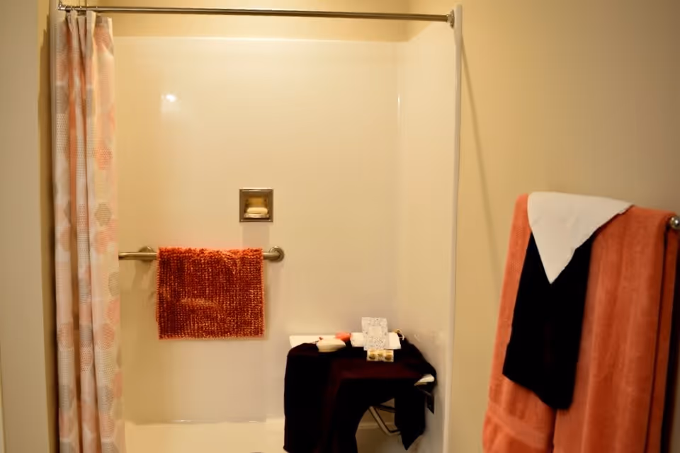A bathroom shower area with a white shower curtain featuring a subtle pattern, a silver shower rod, and a red towel hanging on a grab bar inside the shower. There is a small black table inside the shower holding toiletries and a card. On the right side, a towel rack holds a coral towel with a black towel and a white cloth draped over it. The walls are painted a light beige color.