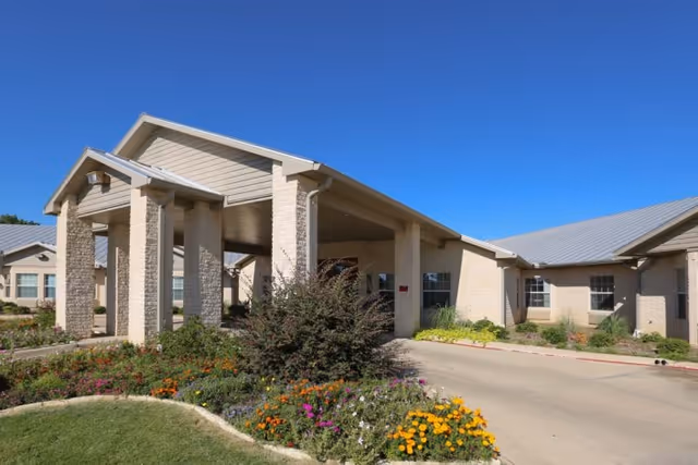 Front entrance of a beige brick senior living facility with a covered porte-cochère and landscaped flowerbeds under a clear blue sky.