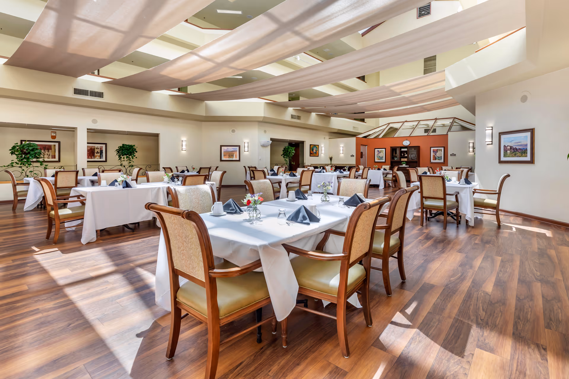 A spacious, well-lit dining room with multiple tables covered in white tablecloths, each set with napkins, cups, and small flower arrangements. The room features wooden floors, comfortable wooden chairs with cushioned seats, and large windows allowing natural light to fill the space. The ceiling has decorative fabric panels and the walls are adorned with framed artwork and plants.