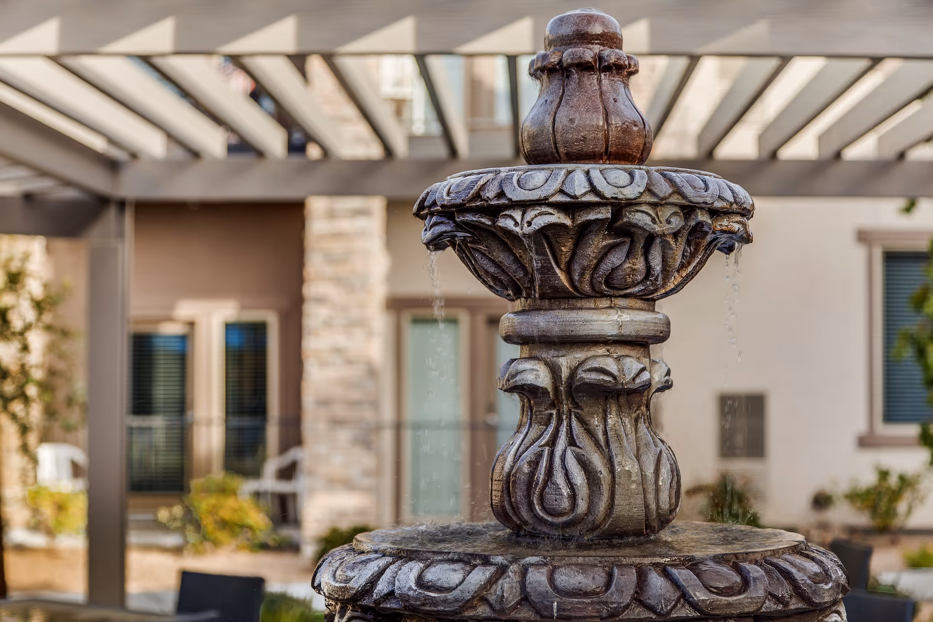 Close-up view of a decorative stone water fountain with water flowing from the top tier, set in an outdoor courtyard area with a pergola overhead and building windows in the background.