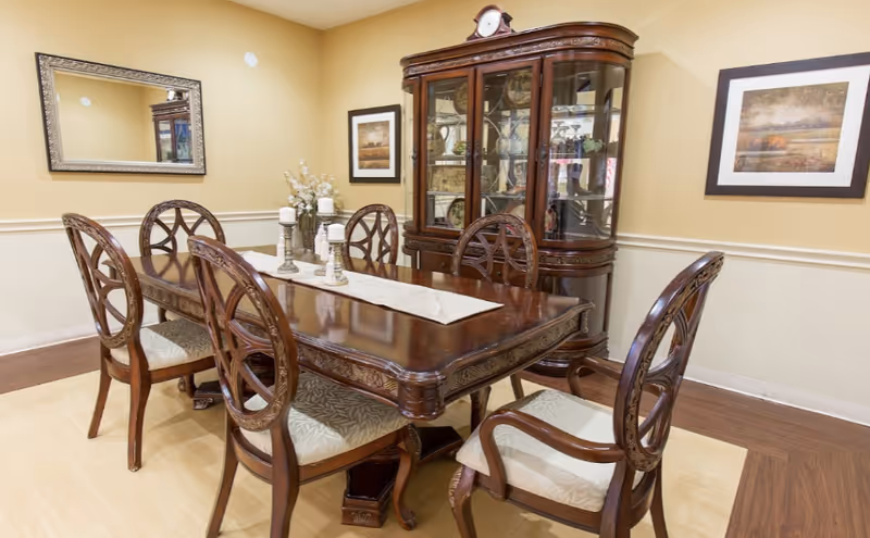 Elegant dining room with a polished wooden table, six ornate chairs, and a glass-front china cabinet against a beige wall.