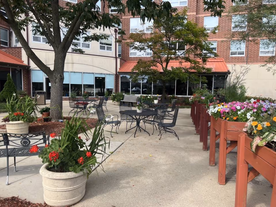 Outdoor patio area at Elison Independent Living of Niles with metal tables and chairs, large potted plants with red flowers, raised wooden flower beds filled with colorful flowers, and trees providing shade. The building's brick and beige exterior with windows is visible in the background.
