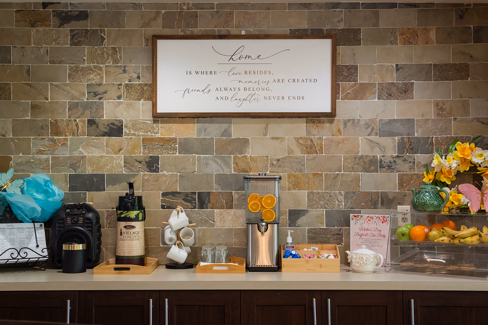 A countertop with various beverage and snack items including a dispenser with orange slices, coffee mugs, a coffee dispenser labeled Village Concepts, a basket with sugar packets, and a fruit display with bananas, apples, and oranges. Above the counter is a framed sign on a stone tile wall that reads 'Home is where love resides, memories are created, friends always belong, and laughter never ends.'
