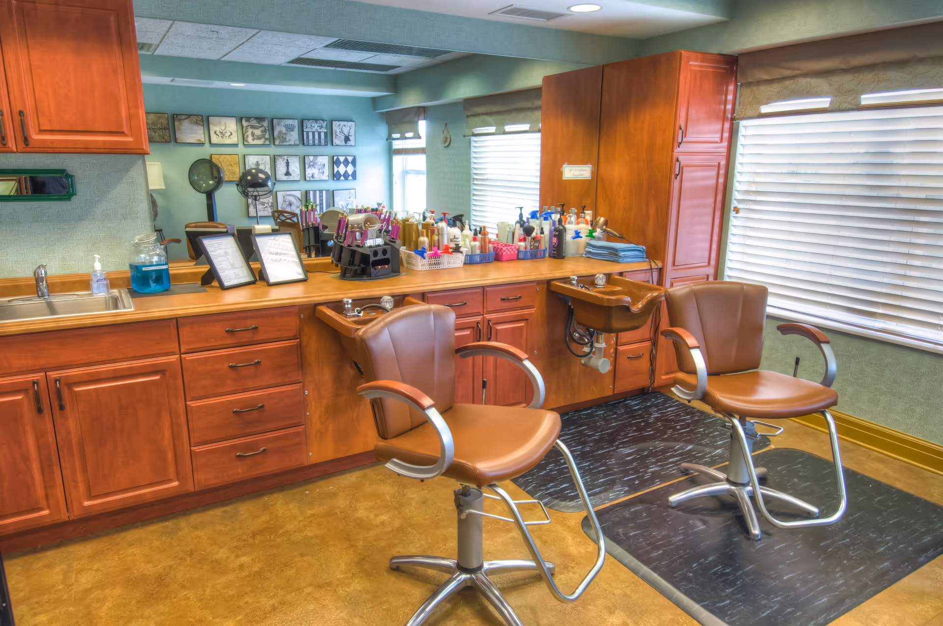 Interior view of a hair salon area with two brown salon chairs in front of two hair washing sinks. The countertop behind the chairs holds various hair care products, spray bottles, and hair styling tools. There is a large mirror on the wall reflecting part of the room, and windows with blinds letting in natural light.