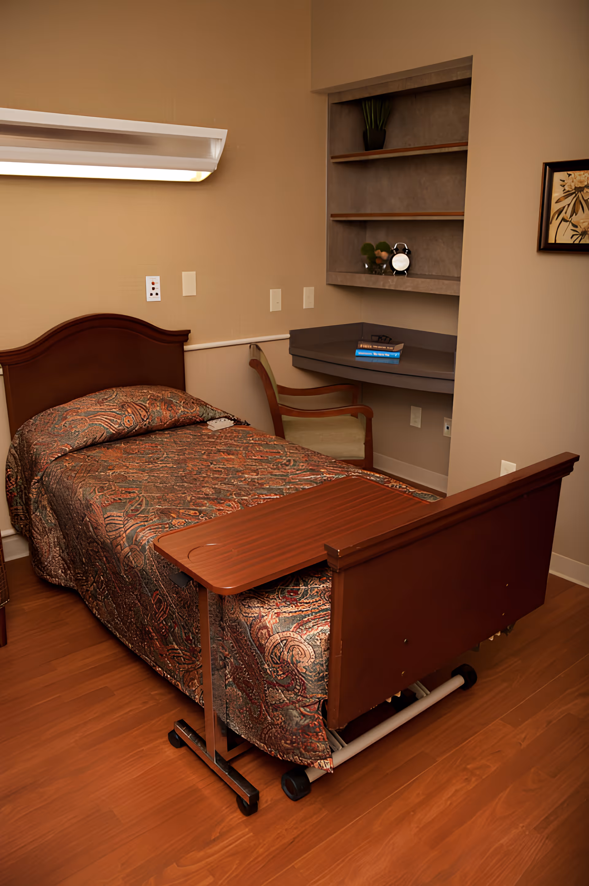 Single-occupancy bedroom with a single bed covered in patterned bedding, an overbed table, a chair, wall shelving, and hardwood floor.