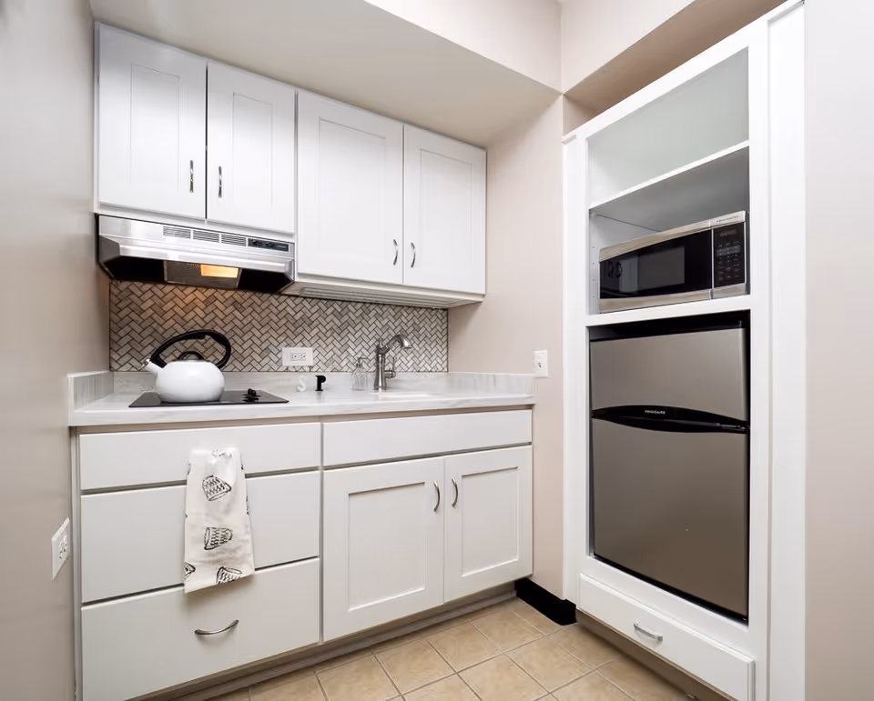 A small kitchen area with white cabinets, a white countertop, a stainless steel range hood, a white kettle on a black stovetop, a sink with a faucet, and a built-in shelf holding a microwave and a mini refrigerator. The backsplash features a herringbone tile pattern.