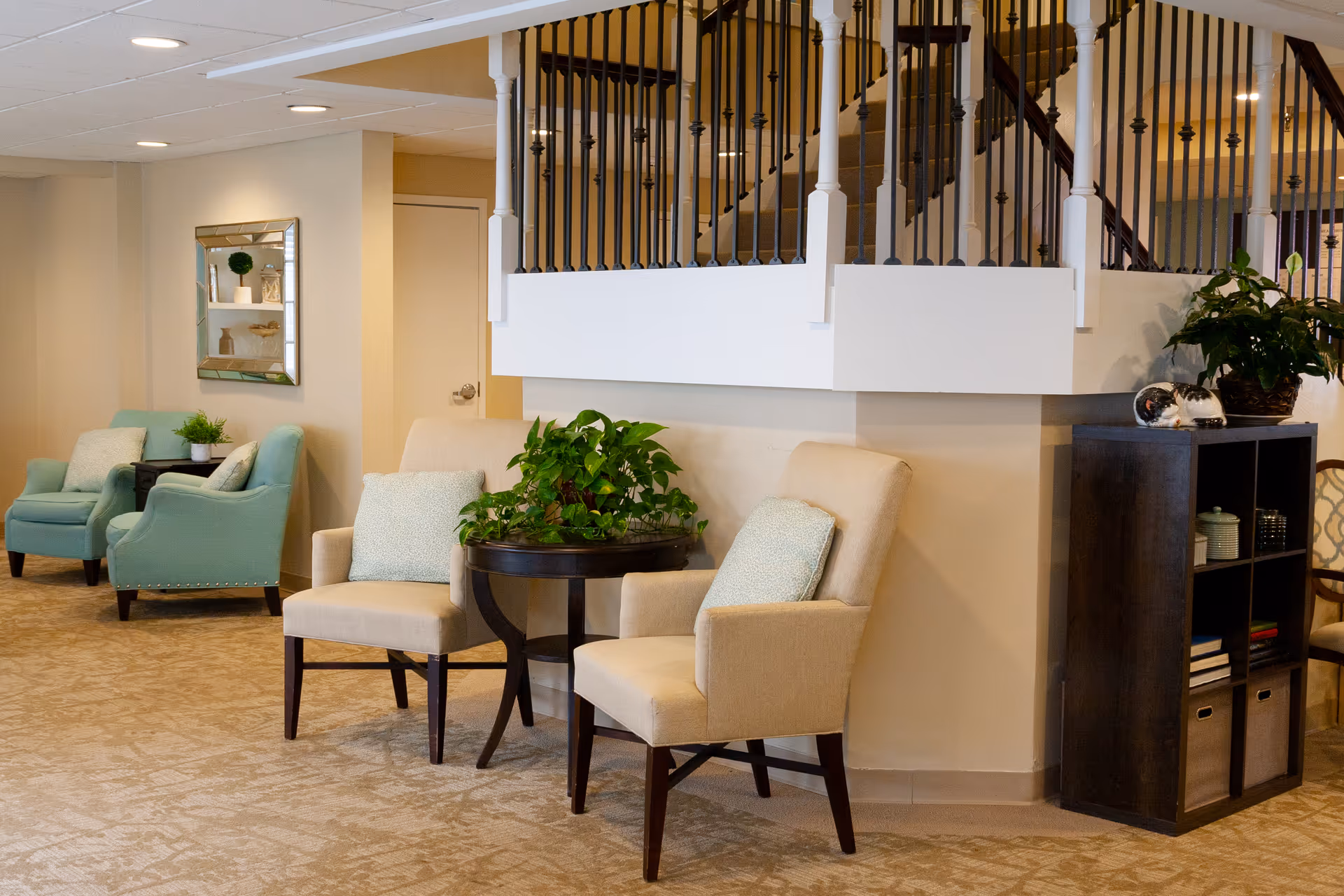 A cozy sitting area inside a senior living facility with beige and light blue armchairs, a small round table with a green plant, a dark wooden shelving unit with decorative items, and a staircase with black metal railings above.