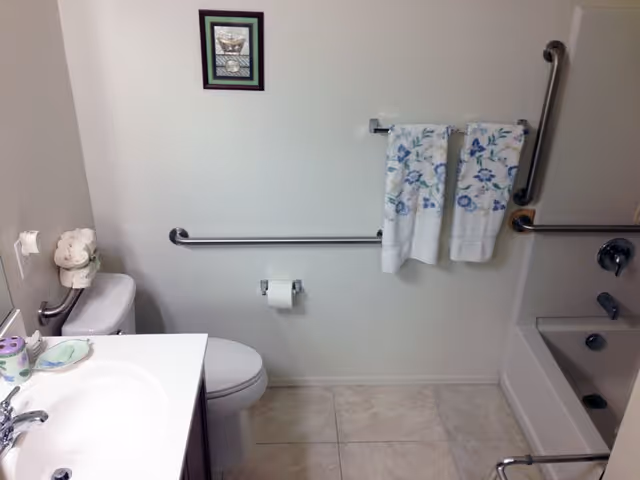 A clean bathroom with a white sink and countertop on the left, a toilet next to it, and a bathtub with grab bars on the right. Two floral-patterned towels hang on a towel rack above the toilet paper holder. A small framed picture is mounted on the wall above the towel rack.