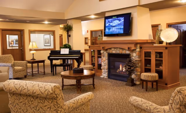 Comfortable common living room with patterned armchairs arranged around a coffee table, a stone-front fireplace with a TV above, and a piano in the background.