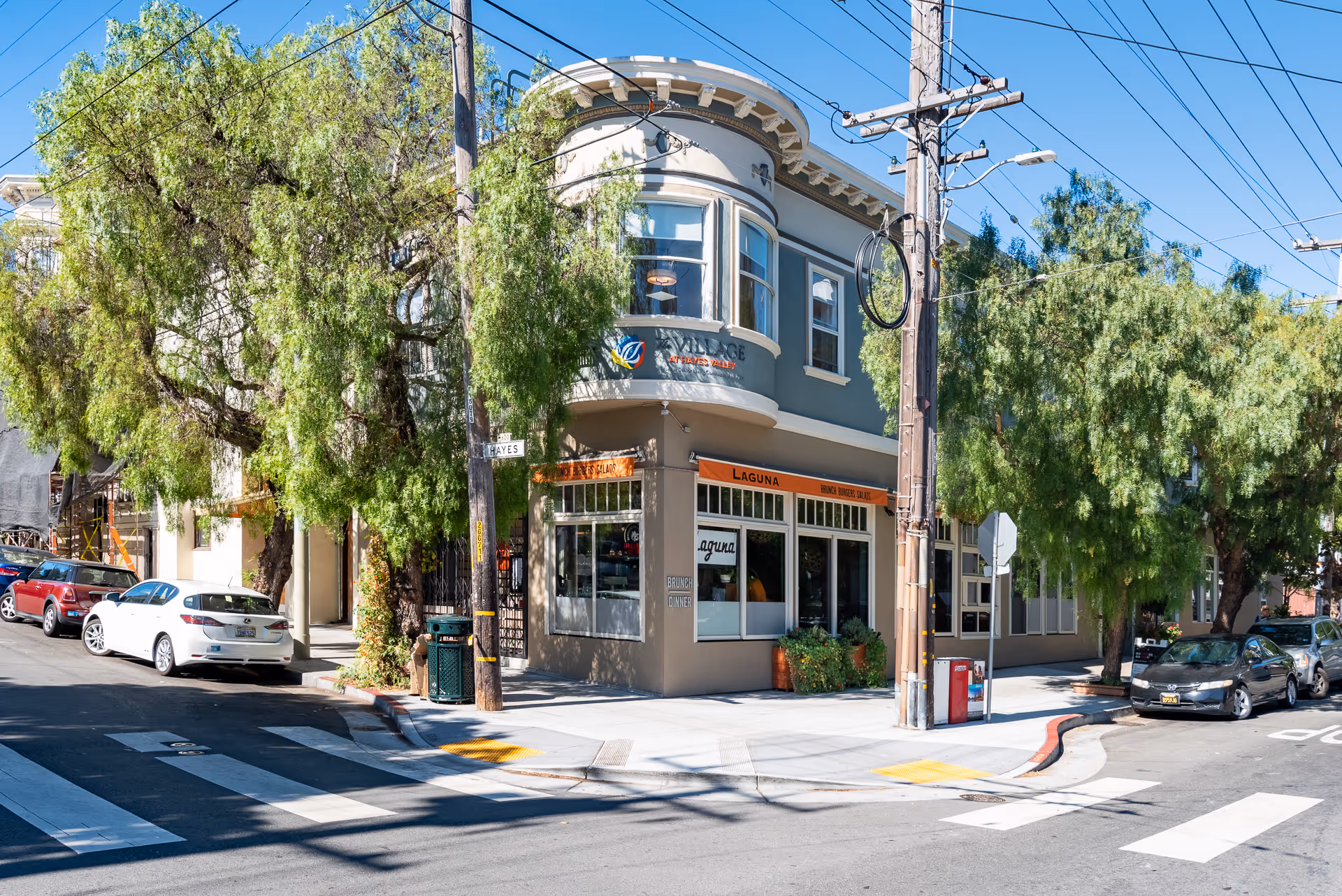 Street corner view of a two-story building with trees lining the sidewalks and several parked cars. The building has a sign that reads 'Village at Hayes Valley' and an orange awning with the word 'Laguna'. Power lines and poles are visible overhead under a clear blue sky.